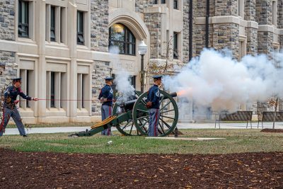 Two cadets stand next to Skipper as a third cadet pulls the lanyard to fire the cannon. Smoke and flame are coming out of the barrel of the cannon as it fires.