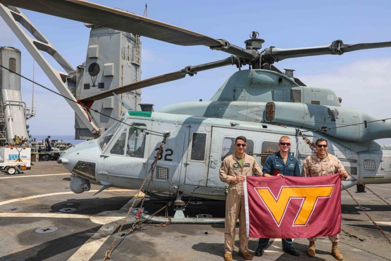 Three servicemembers stand holding a VT flag in front of a helicopter on the deck of a ship. The ocean is in the background.