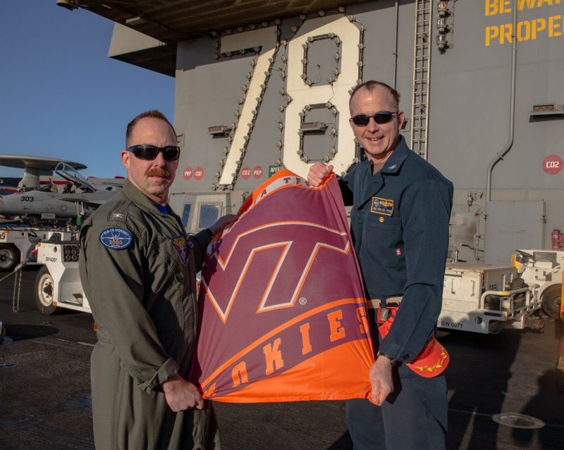Dartez and Von St. Paul stand with a small Virginia Tech flag on the deck of a Navy ship. They are both smiling and wearing military coveralls and sunglasses. 