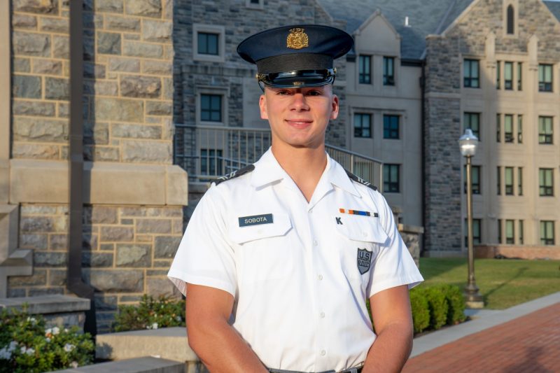 Cadet Sobota stands in his uniform smiling in front of a Hokie stone building. 