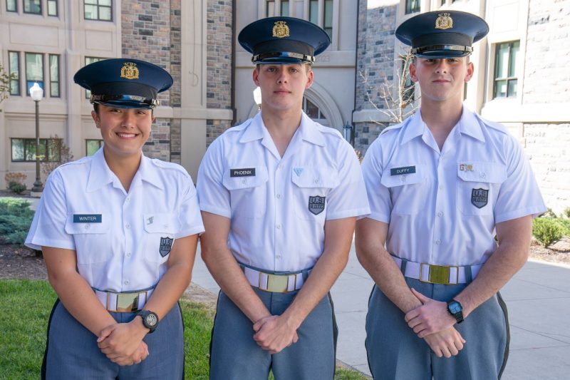 Cadets in uniform stand smiling in front of a Hokie Stone building. 