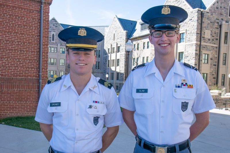 Droney and Elam stand in uniform outside in front of a Hokie stone building and a brick building. They are both smiling. 