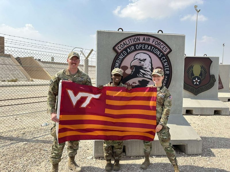 three officers stand in front of a barbed wire fence in the desert while holding a striped VT flag. They are smiling and the sign behind them says “Coalition Air Forces Combined Air Operations Center”. 