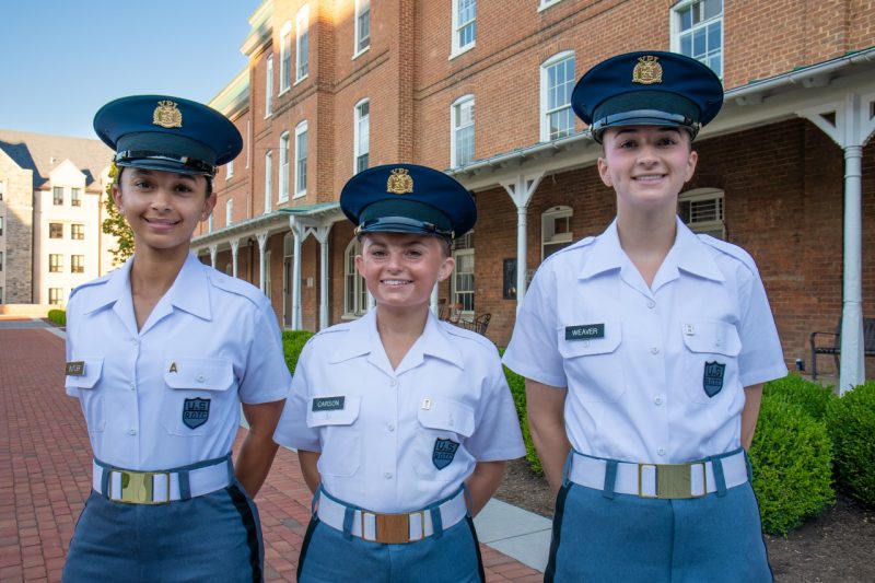 Three women cadets stand in uniform smiling in front of the brick building Lane Hall on a brick sidewalk. 