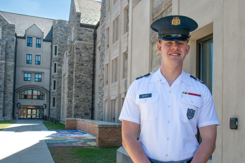 Scott stands in uniform in front of a Hokie stone building while smiling. 
