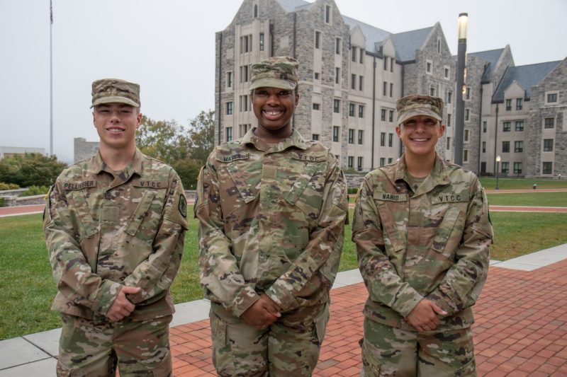 Cadets in uniform stand smiling in front of a Hokie stone buildings. 