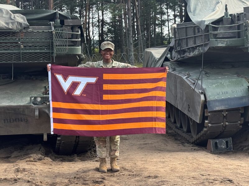A service woman in a camouflage uniform stands in front of two military tanks holding a striped Virginia Tech flag while smiling. 