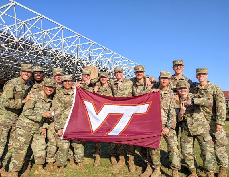 A large group of smiling cadets post with a VT flag outside in camouflage uniforms.