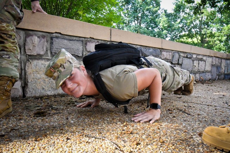 A bright-eyed woman cadet executes a pushup while smiling at the camera. She is in the down position, wearing her backpack and camouflage uniform with a green t-shirt. She looks very strong and proud. 