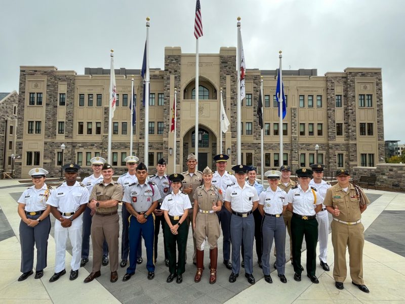 Eighteen cadets in all different military school uniforms stand in front of the CLMS building. Many are smiling and a few have their thumbs up for the picture.