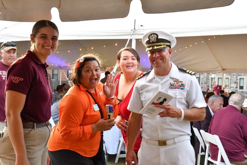 The alumni director stands in his white navy uniform while two other employees smile and wave at the camera. A large event tent is in the background with white lights strung from the ceiling.