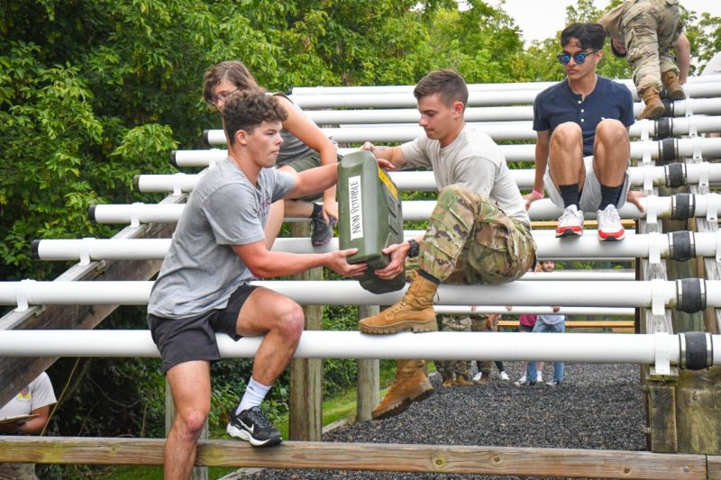two students in the DCTC program, one a cadet and one a civilian student, handle a heavy water jug while sitting on an elevated obstacle on the obstacle course.