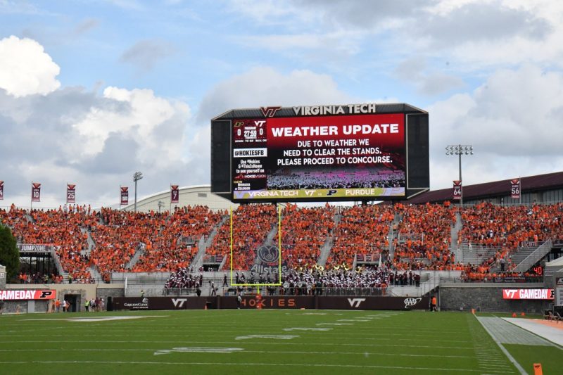The scoreboard at Lane Stadium says "Weather Update Due to Weather We need to clear the stands. Please proceed tot he concourse." Puffy clouds build in the background.
