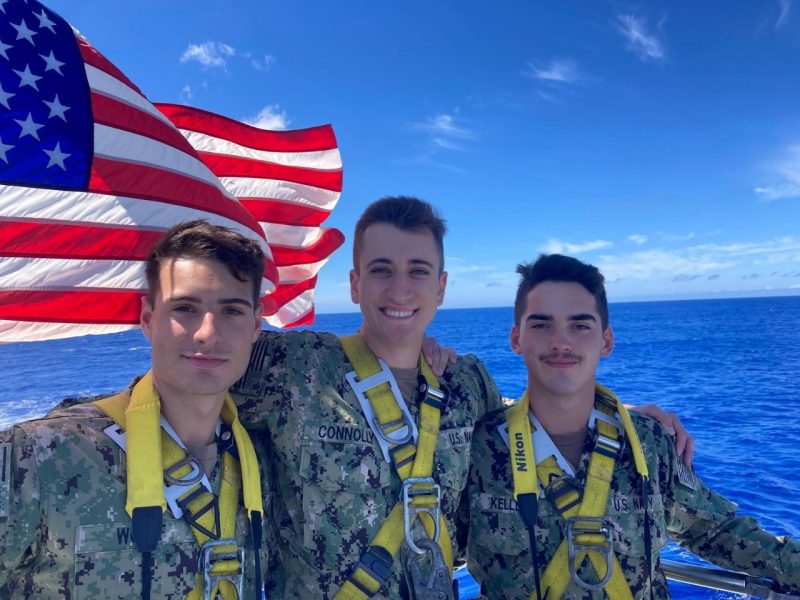 Three midshipmen smile for the camera. The ocean and the American flag are in the background.