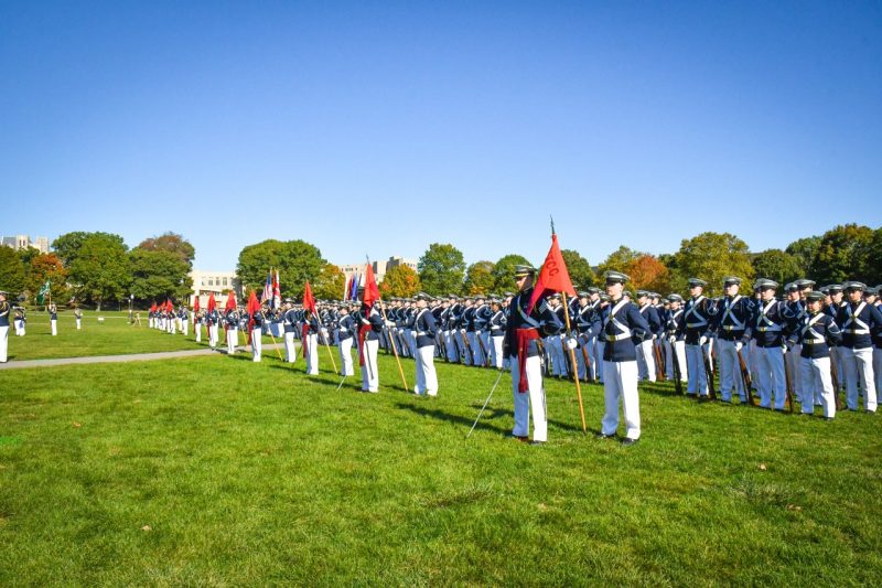 The regiment stands on the drillfield in their dress uniforms for a pass in review. They carry flags for each cadet company and stand at parade rest. The sky is blue and the uniform pants are bright white.