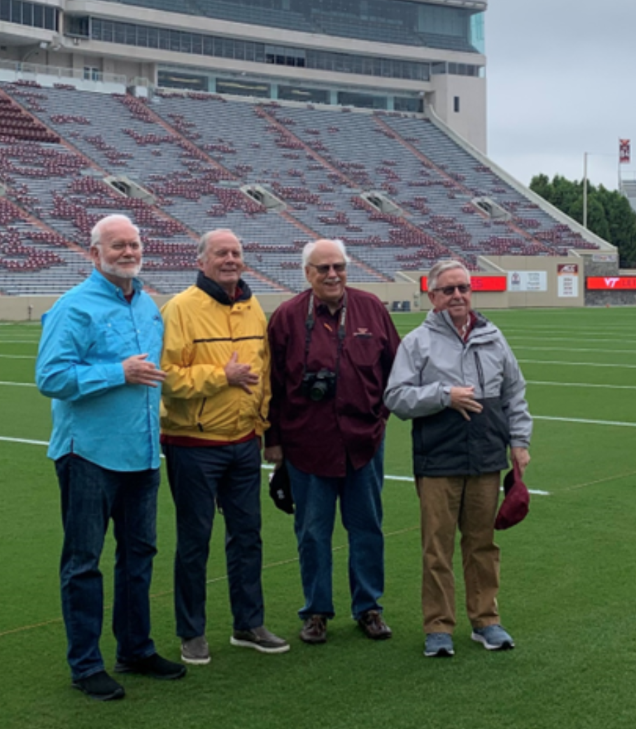 Four smiling men stand on Worsham Field in Lane Stadium.