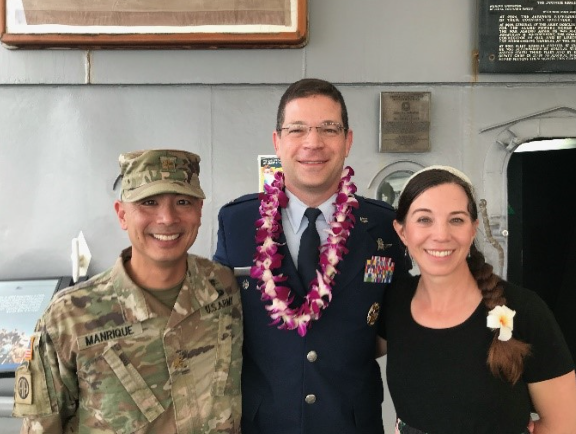 Theising stands in the middle in his dress uniform wearing a lei. Manrique is in his camouflage uniform and Ericka is wearing a black shirt. They are all smiling.