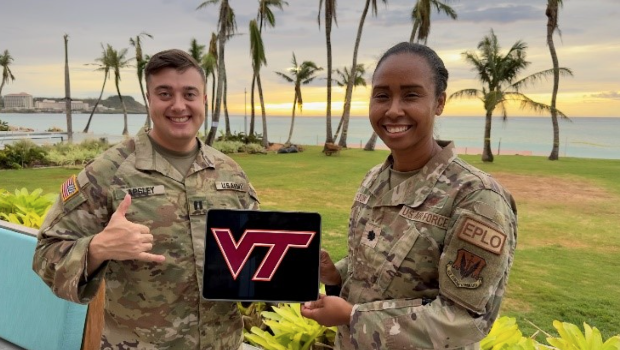 The two stand holding a small VT sign. They are smiling and there are palm trees in the background.