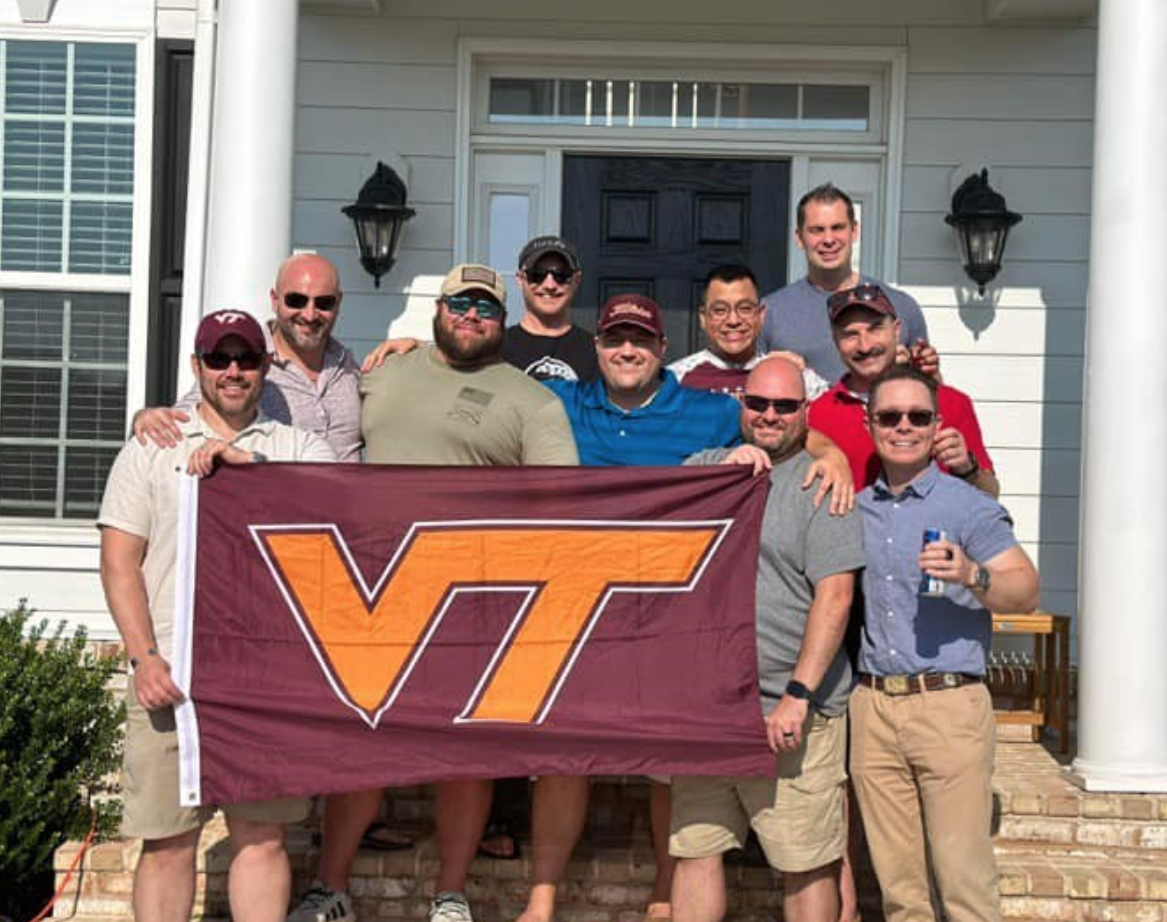 A group of alums stand on the porch of a white building holding a VT flag.