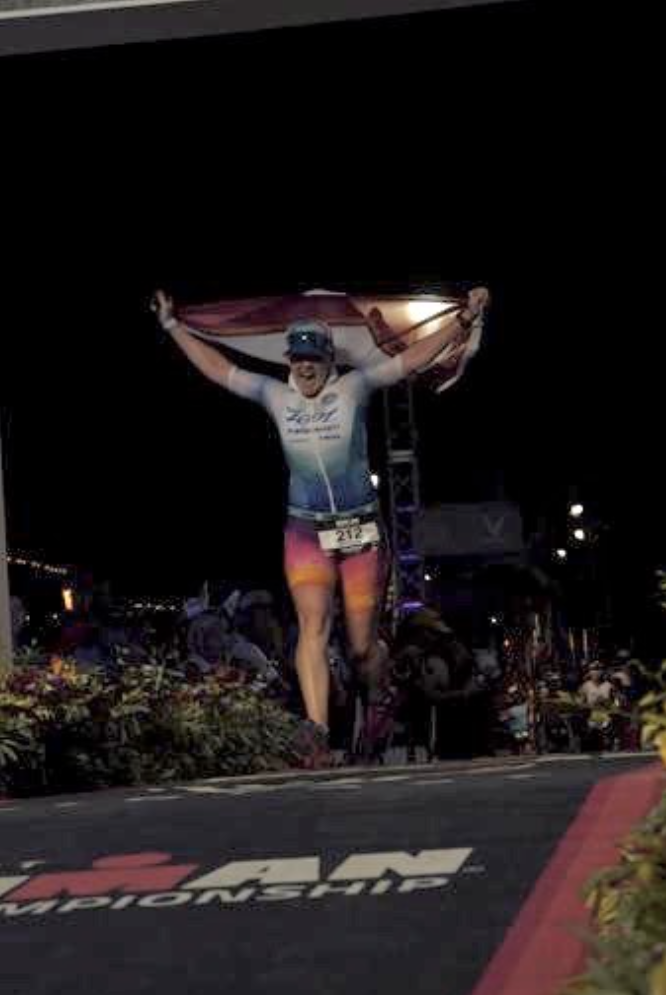 Bethel is holding a flag over her head in both hands while approaching the finish line at night. She is shouting.