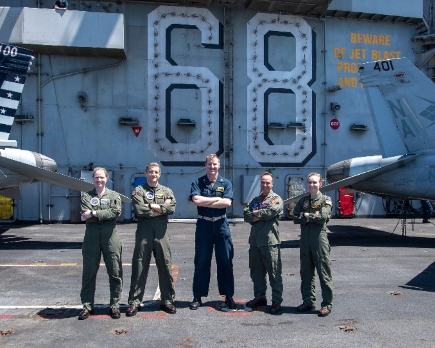 The group stands with arms crossed smiling. All are in military uniforms as they stand in on the flight deck in front of the ships number 68.