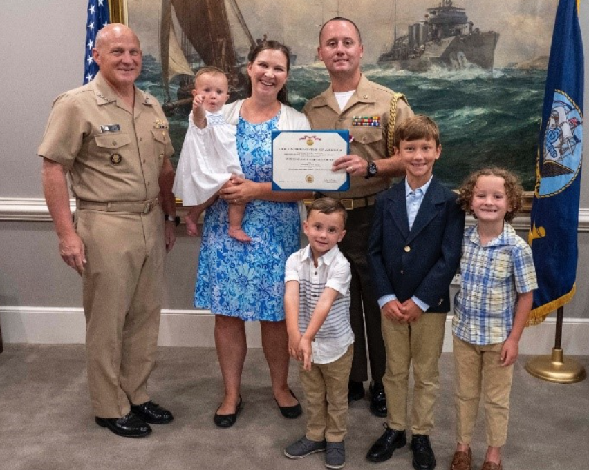 Peterson stands in his uniform smiling with his family. He's holding a certificate and the admiral in uniform stands next to his family.
