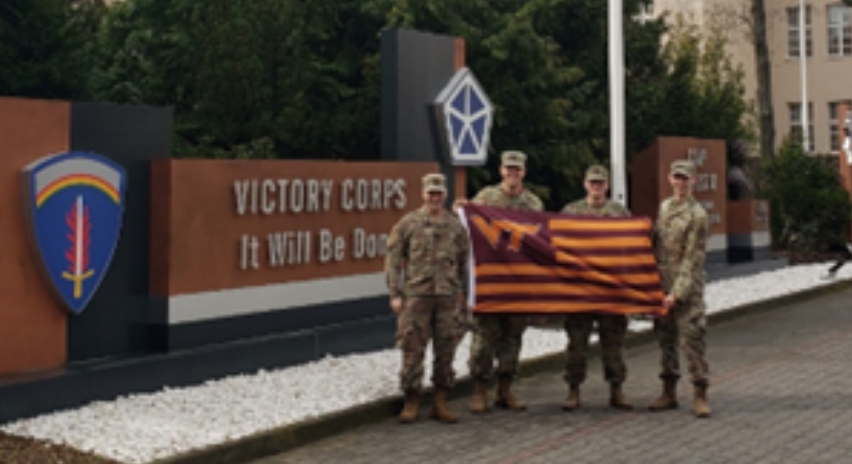 The four stand in front of a large sign reading "Victory Corps it will be done". they are in camouflage holding a striped VT flag.