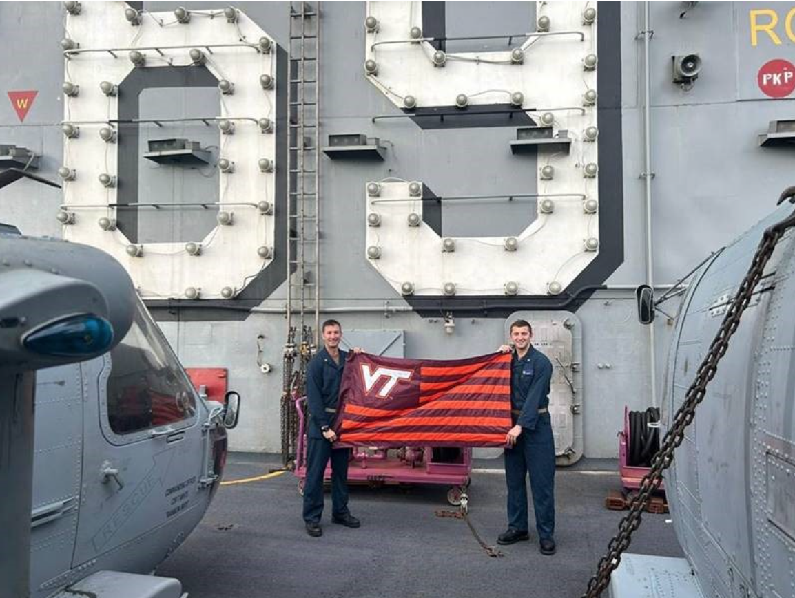 The two stand in blue navy coveralls in front of the ships number 69 on the flight deck holding a striped VT flag.