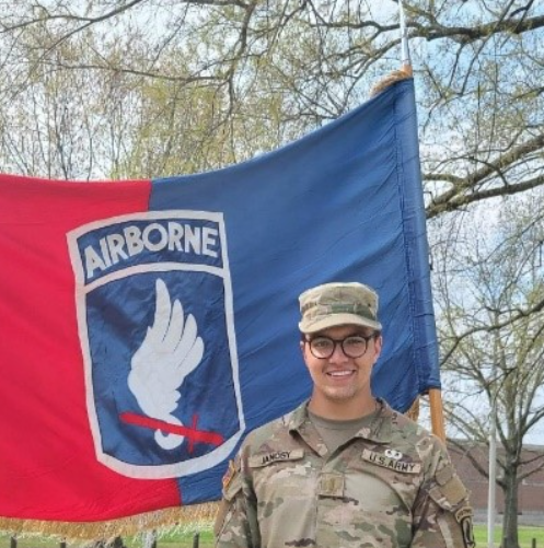 Janosy stands smiling in front of an Airborne flag.