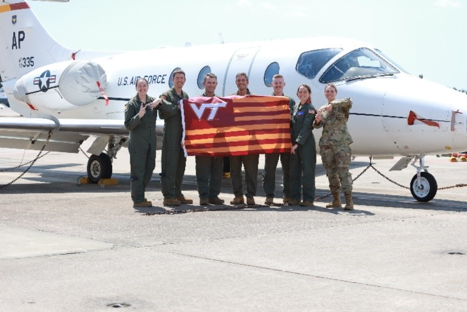 The group stands in front of a US Air Force plane holding a striped VT flag.