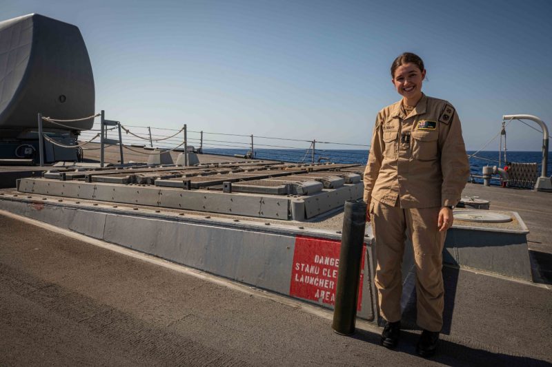 Mason stands next to a missile launcher on a ship smiling. The open ocean is in the background