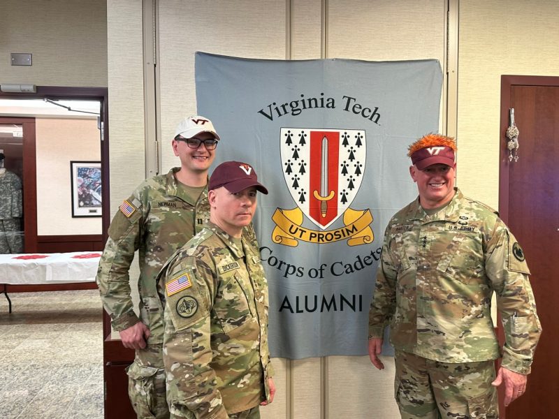 Harter, Newman, and Dickerson stand in uniform in front of a banner with the Corps logo and the words Alumni..