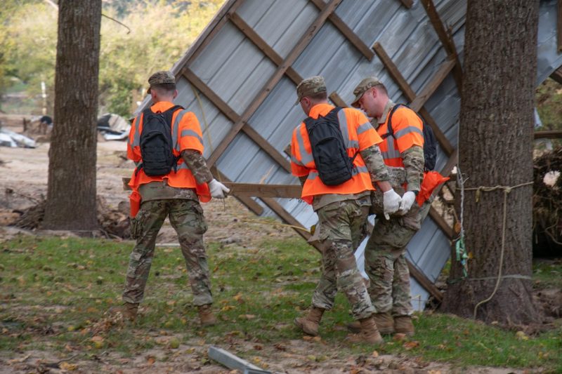 Three cadets carry a fence post with a concrete base near a flipped roof leaning on a tree.