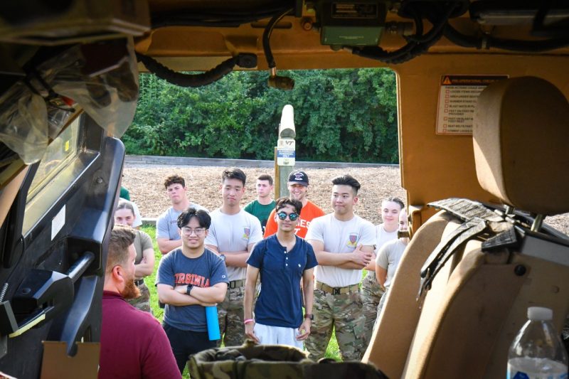 View looking through the window of a military vehicle towards a group of students listening to an instructor speaking about the vehicle.