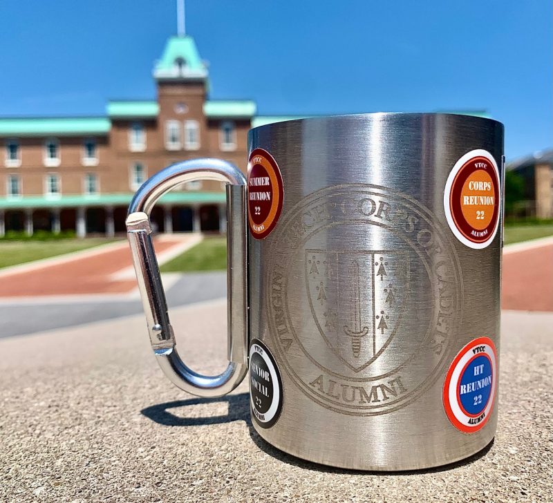 A Corps camping mug sits on Upper Quad with Lane Hall in the background. The mug is adorned with stickers.