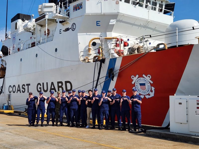 Cadets stand in front of a Coast Guard ship making the VT sign with their hands while smiling.
