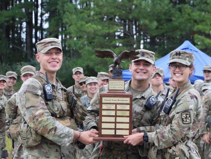 Three cadets hoist a large trophy topped with a bronze eagle while smiling. They are surrounded by smiling cadets in the background.