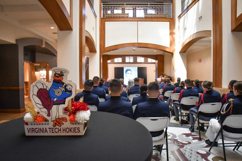 A HokieBird centerpiece sits on a table in the atrium as cadets and alumni watch a speaker at the podium.