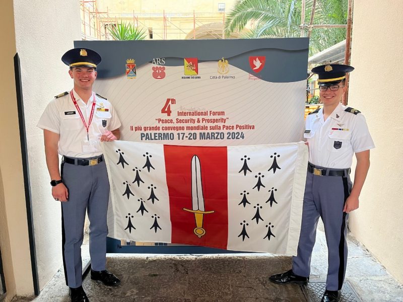 Lerner and Dominique stand with a Corps flag in front of a sign about the conference.