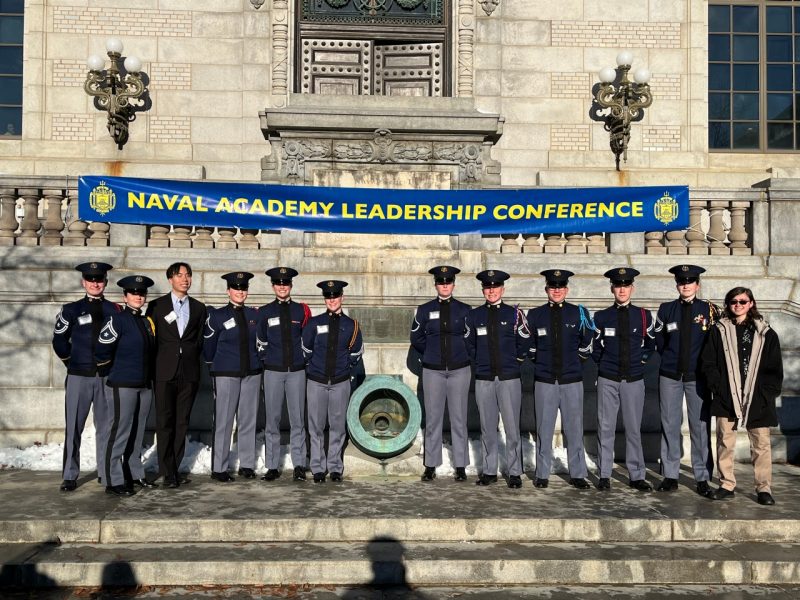 A large group of cadets and students stand and smile for the camera under a banner with the name of the Naval leadership conference.