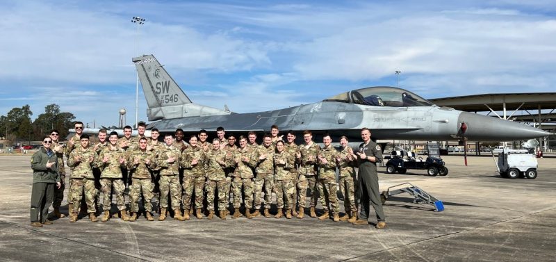 Virginia Tech Air Force ROTC cadets flash the “gunfighter” sign of the 55th Fighter Squadron in front of a military jet.