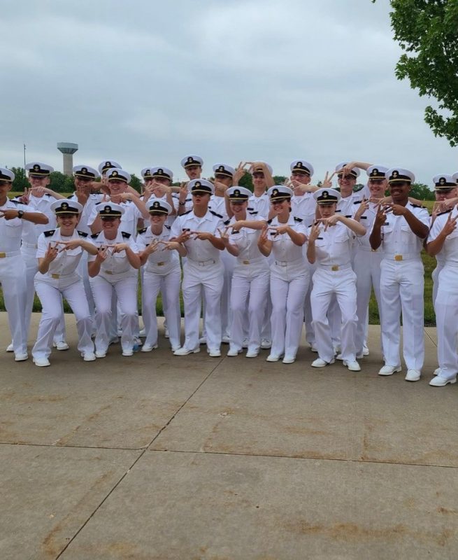 A large group of midshipmen in uniform make the VT sign with their hands while smiling.