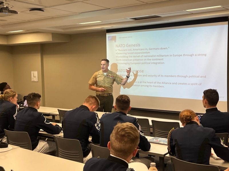 Clark motions in front of a slideshow presentation while speaking to cadets in uniform
