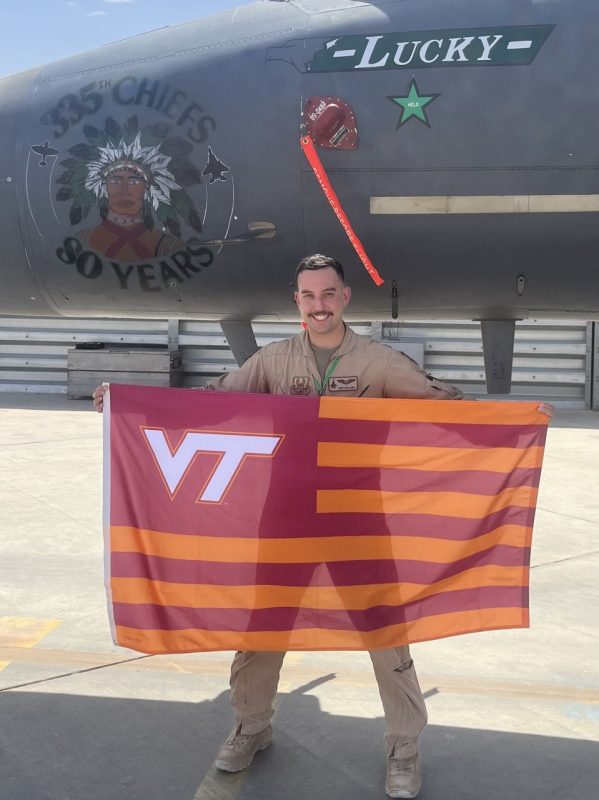 Captain Dimosthenis Doulamis holds a striped Virginia Tech flag while wearing his flight suit. He is smiling and standing in front of a fighter plane on the tarmac.