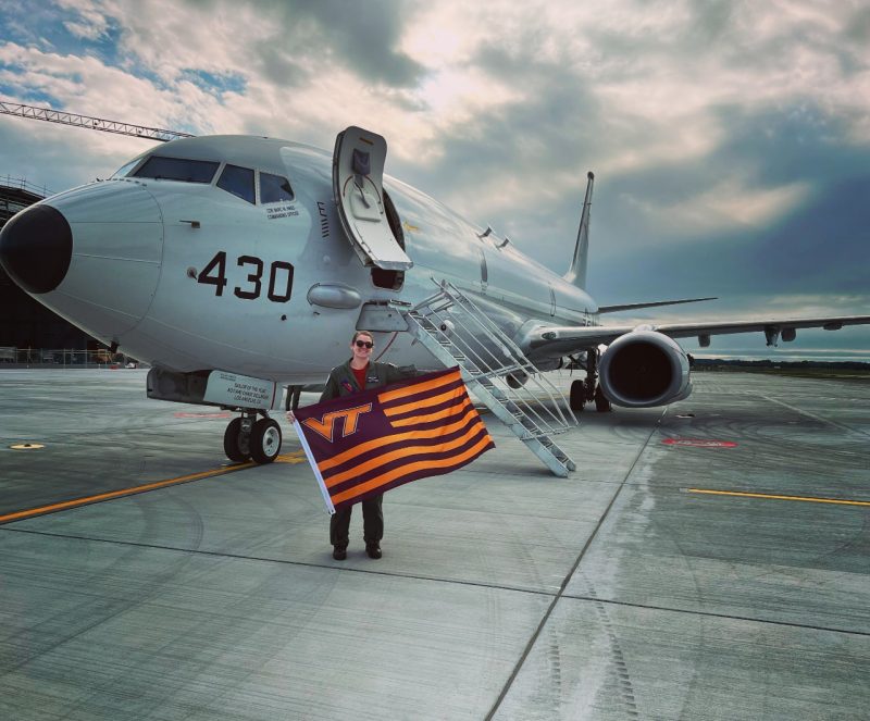 Slaughter stands with a striped Virginia Tech flag in front of a military aircraft. She is smiling and wearing a flight suit.