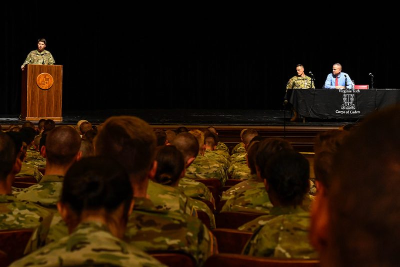 Three men speak from a stage to an audience of cadets.