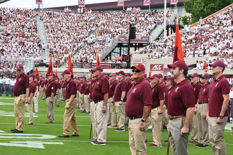 Groups of corps alumni in matching maroon polo shirts and hats stand in formation on a the football field