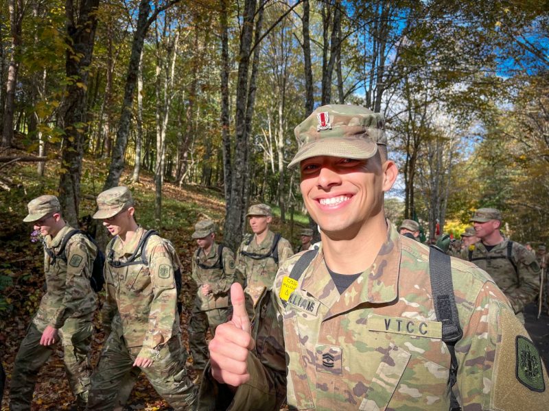 A cadet smiles brilliantly at the camera with a thumbs up. He's in his camouflaged cadet uniform in a wooded area with other cadets behind him.