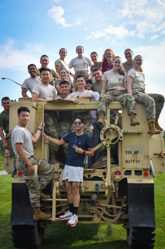 A group of civilian students and cadets all enrolled in DCTC sit and stand on top a military vehicle. Some are in civilian clothes and some are in uniform. They are all smiling.