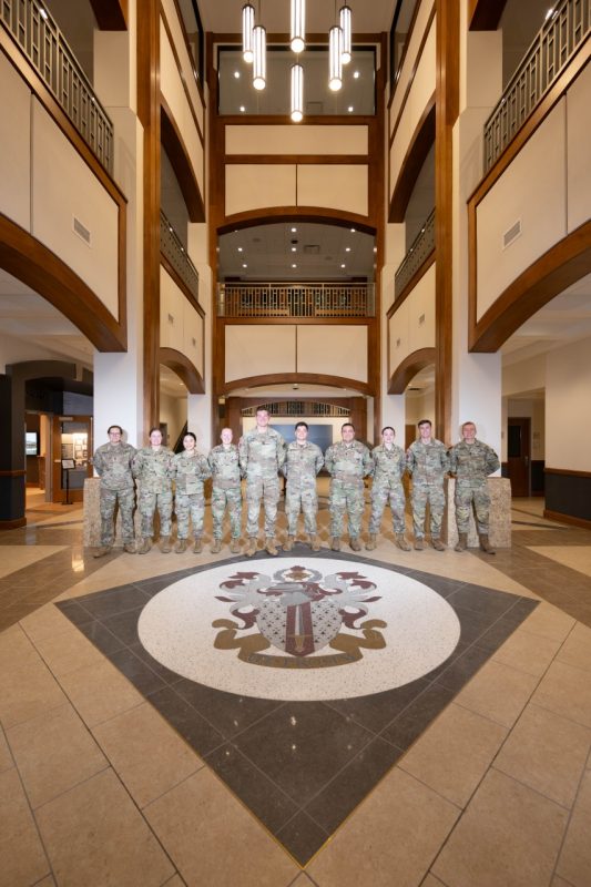 Regimental Staff cadets stand in the atrium of the Corps Leadership and Military Science Building. They are wearing camouflage uniforms and all are smiling.